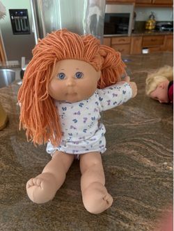 Cabbage Patch Kids doll features distinct orange yarn hair and bright blue eyes, paired with a charming romper decorated with blue and purple flowers