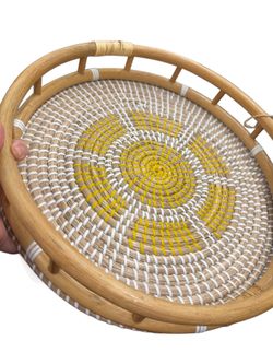 round, hand-woven serving tray features a vibrant yellow and white patterned design on the base and a natural rattan frame with incorporated handles.