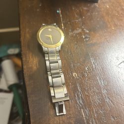 Movado two-tone stainless steel wristwatch with a gold-plated bezel and a black dial. It features the brand's signature single gold-toned dot at the 1