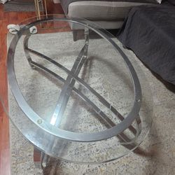 Stylish oval glass coffee table featuring a clear tempered glass top and a sleek chrome crisscross base. The modern design complements most living roo