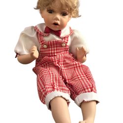 vintage porcelain doll of a young boy with blonde, curly hair, dressed in red and white gingham overalls and a white short-sleeved shirt with a red bo