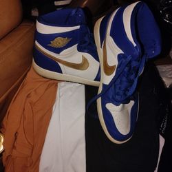 This is a Nike Air Jordan 1 Retro High "Gold Medal" in a white and deep royal blue colorway with a metallic gold Swoosh and Wings logo. 