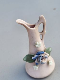 Miniature ceramic or bisque vintage pitcher vase in a pink or pastel finish, decorated with a central blue/purple rose, green leaves, and small applie
