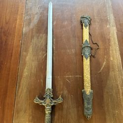 Decorative medieval-style short sword with ornate scabbard. The sword features a detailed bronze-tone handle with red gem accents and intricate design