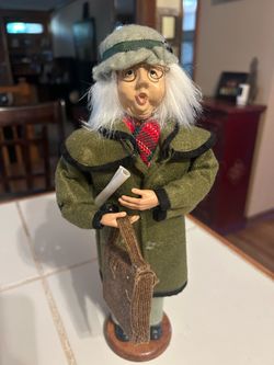 Byers' Choice Caroler figurine of an older woman wearing a green coat, wire-rimmed glasses, and a hat, holding a scroll and a corduroy handbag