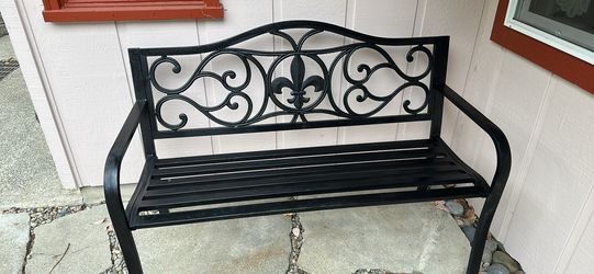 Sturdy metal garden bench is black with a decorative fleur-de-lis and scrollwork backrest. This outdoor seat has a slatted seat and curved armrests.  