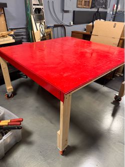 Custom-built work table features a vibrant red painted top supported by raw pine legs equipped with caster wheels - 60” x 60” x 37”