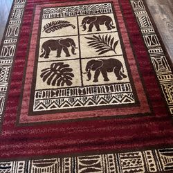 Brand new condition Burgundy rectangular area rug has central panel with elephant and tropical leaf motifs. Border features cream and black tribal geo