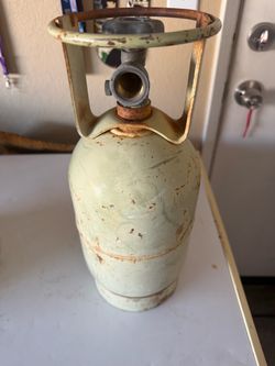 Older weathered vertical gas cylinder, likely a vintage or standard portable steel propane tank with a ring collar handle and POL valve