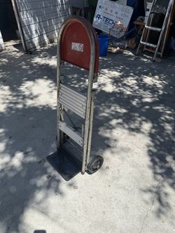 vintage two-step aluminum household folding ladder and utility cart with a red top tray, handrail loop, and wheels at the base.
