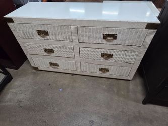 This chest of drawers is a vintage piece—featuring Hollywood Regency or Campaign style—made of white wicker with brass hardware and corner fittings.