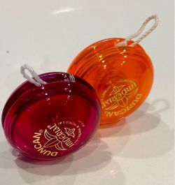Two Duncan Imperial yo-yos in transparent pink and transparent orange. Both have gold logo imprints and white string attached.