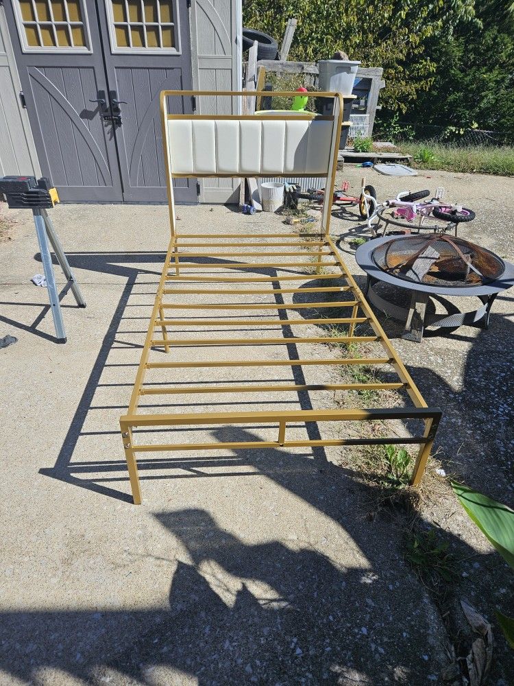 This is a gold-colored metal bed frame with a white upholstered headboard.
The bed frame is a twin