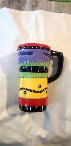 Vibrant multi-colored ceramic travel mug has a hand-painted style design. The exterior has horizontal bands of purple, green, yellow, and red with rai