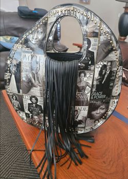 Stylish circle tote with magazine-cover collage print and long black fringe. Bold gold stud accents add luxe detail. Lightweight and eye-catching for 