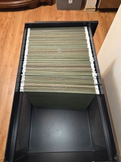 Black metal file cabinet with a flip-top lid and front door, loaded with green hanging file folders. Features a mesh side panel, open bottom shelf, an