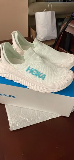 The Hoka Restore TC slip-on in the Frost/Cloudless colorway features a breathable, sock-like mesh upper with bright blue branding. Men 12