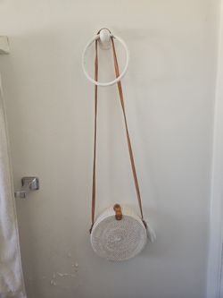 This is a white rattan round crossbody bag with a brown leather strap and closure.