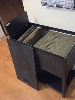 Black metal file cabinet with a flip-top lid and front door, loaded with green hanging file folders. Features a mesh side panel, open bottom shelf, an
