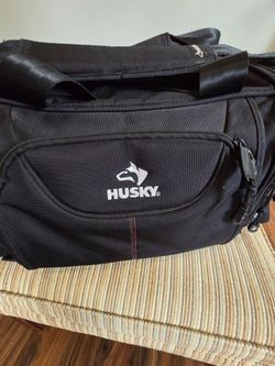 black Husky Duffle Bag made of heavy-duty, water-resistant material with dual carrying handles and a front zippered pocket bearing the white H
