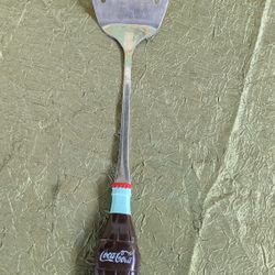 Coca-Cola branded collectible kitchen utensil, specifically a slotted spatula with a handle shaped like a classic Coca-Cola bottle.
Material: Stainles