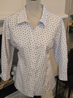 Crisp white button-down blouse patterned with black polka dots. Fitted silhouette with a classic collar. Long sleeves and front button placket. Versat