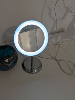 Round tabletop vanity mirror with a glowing LED ring light and chrome pedestal base with touch button control 3x magnification