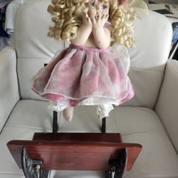 Blonde curly-haired porcelain doll in pink dress on a wooden rocking chair.