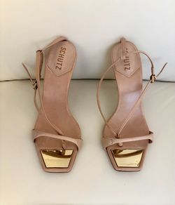 The Schutz Vikki heeled sandals are made of beige leather. Their distinctive square toe incorporates a shiny gold metal plate on the insole.