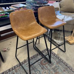 Pair of modern counter height bar stools with whiskey brown faux leather seats and black metal legs - 26” T