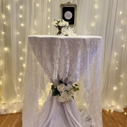 Elegant Wedding/Event Cocktail Table Setup  White table cover with delicate lace overlay Stylish floral arrangement with white roses & greenery 