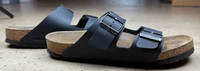 Classic Birkenstock Arizona sandals with black synthetic straps and adjustable metal buckles. The footbed is made of cork and latex with a suede linin
