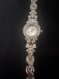 Elegant silver-tone quartz watch has a round face with a rhinestone-encrusted bezel. The decorative link bracelet is adornedi with clear rhinestones t