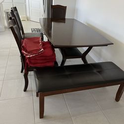 Dark brown dining table set with 4 chairs and a bench. Chairs and bench have black leather seats. Includes 4 red seat cushions and 2 red tablecloths. 
