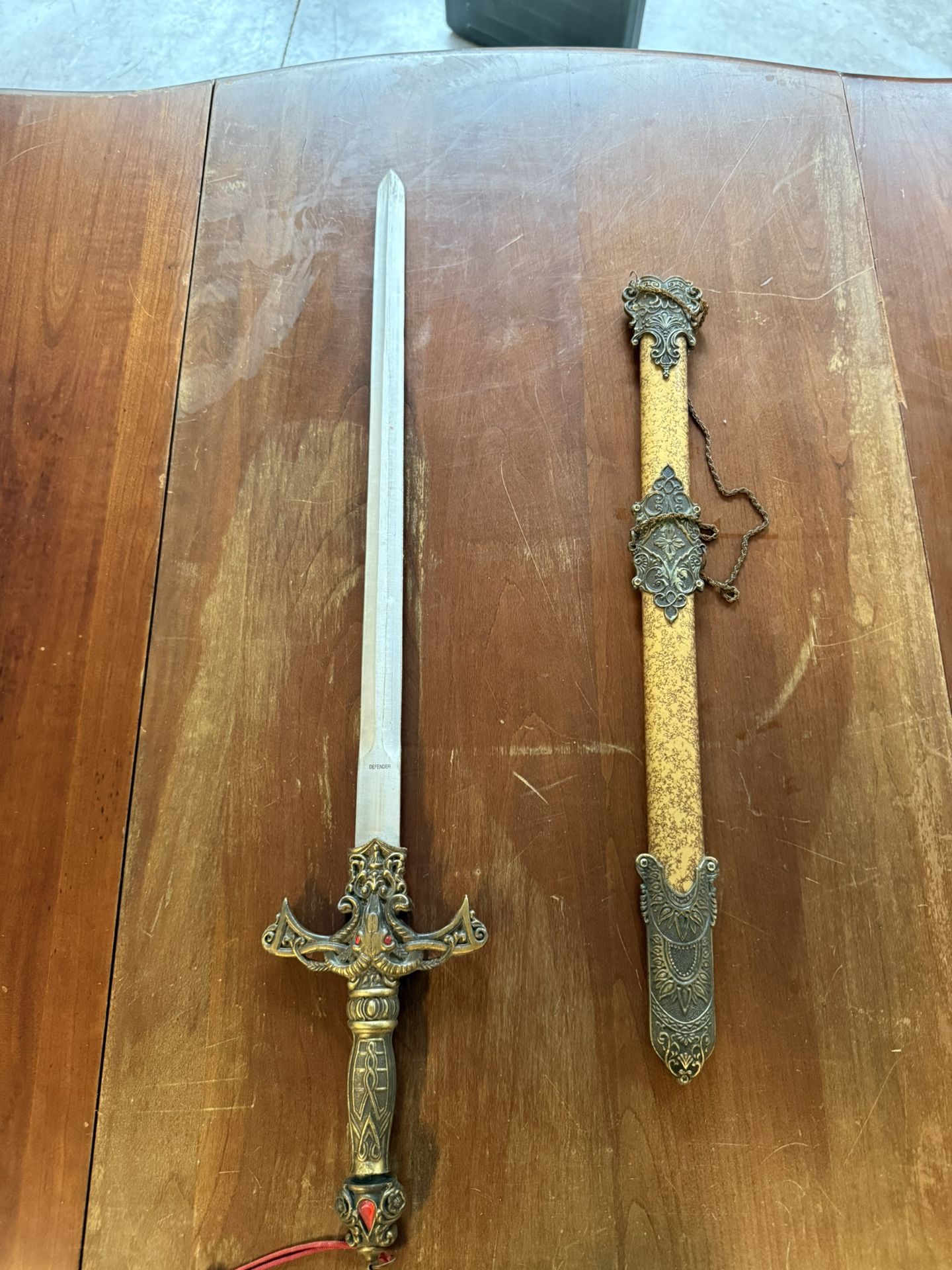 Decorative medieval-style short sword with ornate scabbard. The sword features a detailed bronze-tone handle with red gem accents and intricate design