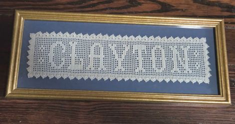 framed, long, rectangular piece of handmade filet crochet lace with the name "CLAYTON".