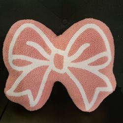 Fuzzy pink bow-shaped decorative pillow with white detailing. 12" H x 15.5" W x 4" D