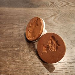 2 vintage unglazed terra cotta cookie stamps featuring embossed animal designs, complete with a round ceramic base and a small white handle on the bac
