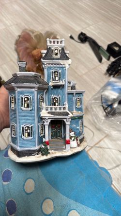 This is a blue Victorian-style house from the Holiday Time Christmas village collection, featuring a small snowman standing near the front door.