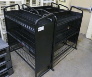 Black powder-coated metal retail display unit with multiple wire shelves on all sides and push-handle rails along the top. Freestanding island-style f