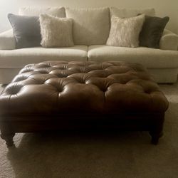 Beautiful faux leather button tufted ottoman coffee table with aged bronze nail head trim. Very good used condition. Solid wood base with bun style fe