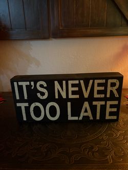 Rustic black wooden box sign distressed white block lettering that reads "It's Never Too Late