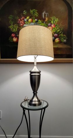 Fangio Lighting metal table lamp features a brushed steel base with a classic chalice design. It has an ivory-white cylindrical linen shade.