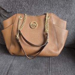 Stylish Michael Kors tote bag is made of brown saffiano leather. It has gold-tone chain and leather shoulder straps. Interior is lined with signature 