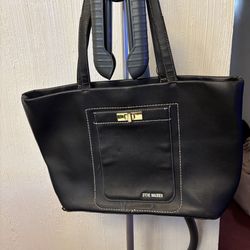 A sleek and stylish black tote bag from Steve Madden featuring a front pocket with a gold-tone clasp detail, white stitching accents, and a spacious i
