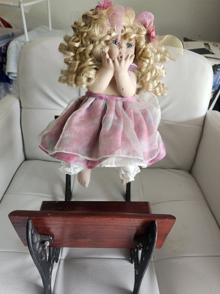 Blonde curly-haired porcelain doll in pink dress on a wooden rocking chair.