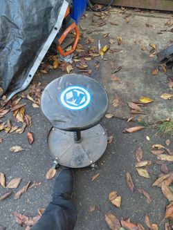 rolling shop/mechanic stool with a black padded seat, a pneumatic height-adjustment lever, a built-in lower tool tray, and casters