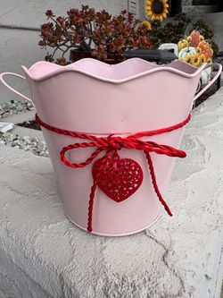 light pink, scalloped-rim metal bucket decorated with a red twine bow and a dangling hollow red heart charm