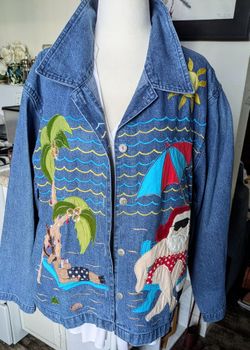 Casual blue denim jacket featuring vibrant embroidered beach scene. Bold stitched sail, palm trees, sun, and Santa lounging motifs. Button-front style