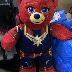 The image shows a Build-A-Bear Captain Marvel plush toy. Key details include:  It is a red, sparkly bear wearing a Captain Marvel costume.  The costum